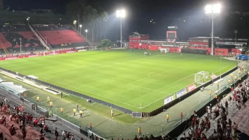 Torcedor rubro-negro do Vitória com a camisa do clube erguendo os braços em momento de gol no Barradão, com a arquibancada ao fundo lotada e iluminada